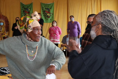 One Black elder woman with a beautiful head piece conversing with an elder Black man with long grey locs and a white beard